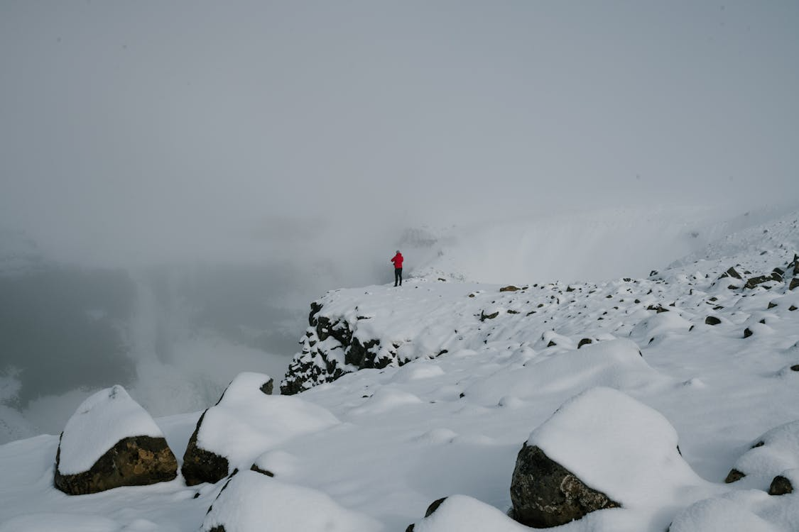 A lone person in a red jacket stands on a snowy cliff amid rocks and dense fog in a cold, mountainous landscape.