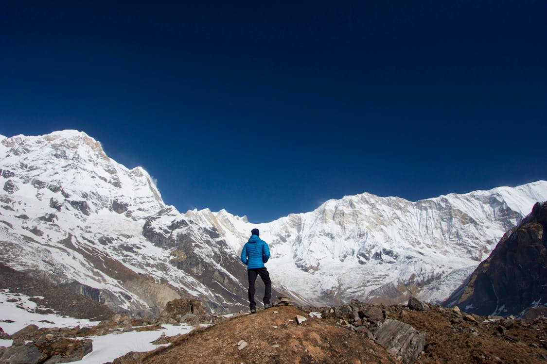 A lone hiker stands on a rocky ridge, gazing at vast snow-covered mountains under a deep blue sky.
