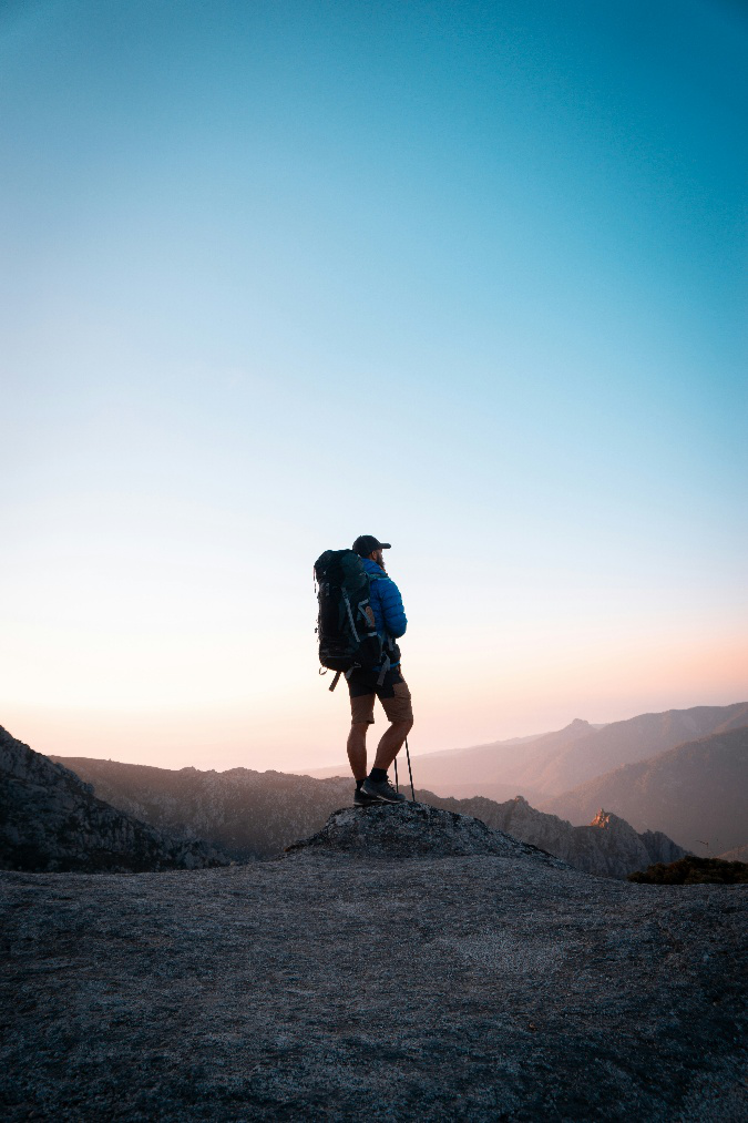 Hiker standing on a rocky overlook with a backpack while surveying a mountainous landscape at sunrise or sunset.