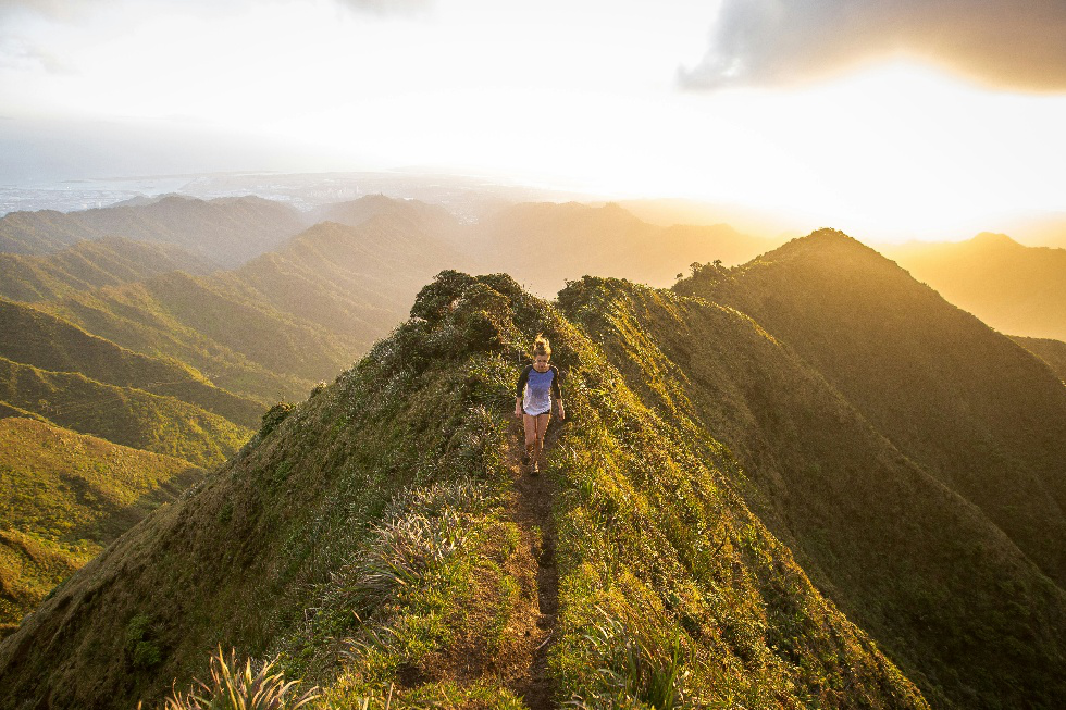 Hiker walking along a narrow mountain ridge trail surrounded by expansive views and golden sunlight.