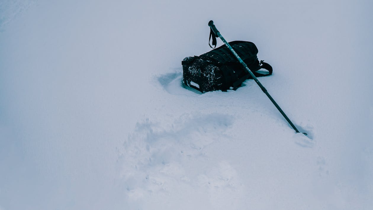 Weather-resistant ski touring bag resting in deep snow with trekking pole during a backcountry winter route.